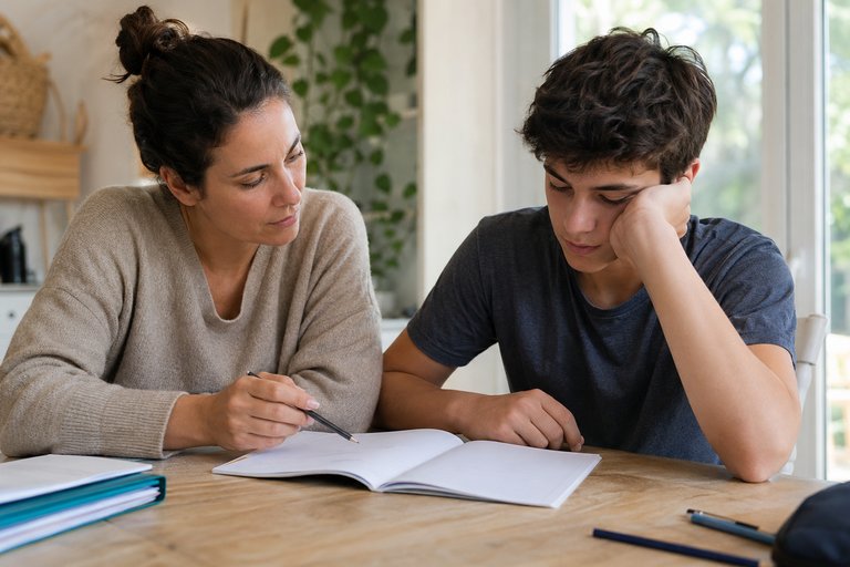 Une adulte assise à côté d'un adolescent découragé devant son cahier, elle l'aide à identifier précisément ce qui bloque.
